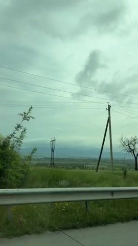 From the passenger seat of a vehicle moving along a highway, the viewer sees a slightly overcast sky. The vehicle passes a guard rail with green vegetation behind it, including some yellow flowering shrubs. Power lines stretch across the frame. In the distance, a flat landscape is visible. A black car is briefly seen on the side of the road.