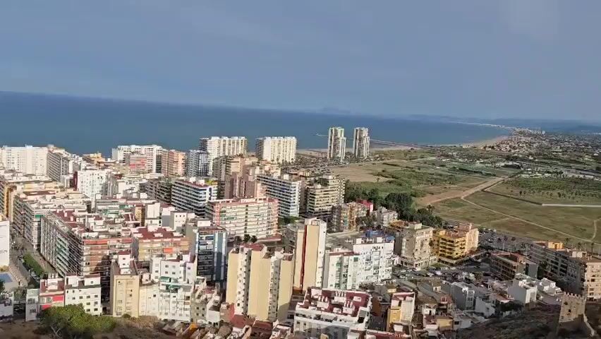 A panoramic drone shot captures a sprawling coastal city, with a vast expanse of blue ocean serving as the backdrop. The city is densely packed with buildings of varying heights and architectural styles, predominantly residential apartments. The structures are a mix of white, beige, and pastel colors, some with colorful balconies. Among the cluster of buildings, two distinct tall, modern white skyscrapers stand out in the distance, closer to the coastline. Beyond the city, a strip of sandy beach is visible, with some vegetation and undeveloped land to the right of the frame. The sky is partly cloudy with patches of blue. In the foreground, a partially visible, weathered, cylindrical stone structure, possibly a historical ruin or tower, is present on a hillside, along with some sparse green vegetation. A black metal railing partially obstructs the view from the lower left quadrant of the frame. A blue trash bin is also visible near the railing. The drone appears to be moving laterally across the scene, offering a sweeping view of the cityscape and the sea.