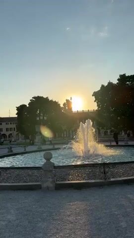 The video opens with a scenic view of a fountain in a plaza, with the setting sun casting a warm glow. The golden hour light illuminates the water, creating a shimmering effect. In the background, silhouetted trees and a building with arched windows stand against a clear sky. A few people can be seen relaxing near the fountain.

The scene then shifts to a wider shot of the plaza, showing the gravel ground, more trees, and lampposts. A couple, a man in a suit and a woman in a white dress, are walking in the distance.

The perspective changes again, bringing the viewer closer to a young woman posing for the camera. She is wearing a sparkly white top and white pants, with her dark hair framing her face. She has a playful expression, puckering her lips and striking a pose. In the background, the same couple from earlier is visible, as well as other people enjoying the park. The setting appears to be a public square or park in Padua, Italy, given the architectural style of the buildings and the overall ambiance of a European city. The time of day is clearly late afternoon or early evening, indicated by the low angle of the sun and the long shadows. The weather is pleasant, with a clear sky and warm lighting.