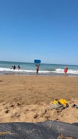 A man is walking out of the ocean and onto a sandy beach carrying a blue and yellow boogie board above his head. He has tattoos on his chest, arms, and legs. The water is choppy with small waves. In the background, other people are in the water. The sky is clear and blue. In the foreground, partially obscured by shadow, are some flip-flops and a yellow bag on the sand.
