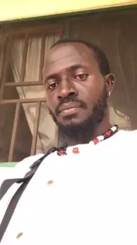 The video shows a man with dark skin, a black beard, and a nose piercing, looking directly at the camera. He wears a white shirt and a necklace with red, white, and black beads. A black strap is visible across his chest, suggesting he might be wearing a bag. Behind him, there's a window with a brown frame and white curtains. At one point, the man briefly sticks his tongue out and touches his chest with his finger.