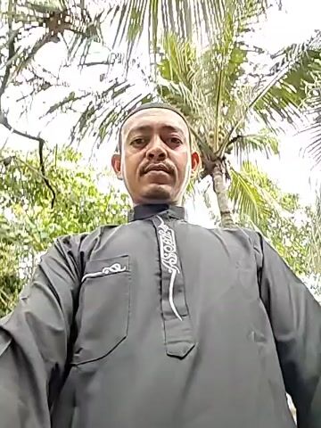 The video shows a man standing outside. He is wearing a black, long-sleeved garment with white embroidery. He is wearing a cap on his head. He is looking directly at the camera. Behind him are palm trees. The sky is overcast.