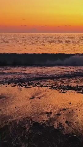 The video captures a close-up view of waves gently crashing onto a sandy beach during sunset. The sky is a vibrant gradient of orange and yellow. The water is a deep blue with foamy white crests as the waves roll in. The wet sand on the beach reflects the warm hues of the sunset, creating a shimmering effect. Small dark pebbles are scattered across the sand, and the water recedes, leaving wet streaks on the shore. The overall scene is peaceful and serene, emphasizing the natural beauty of the coastal environment at dusk.