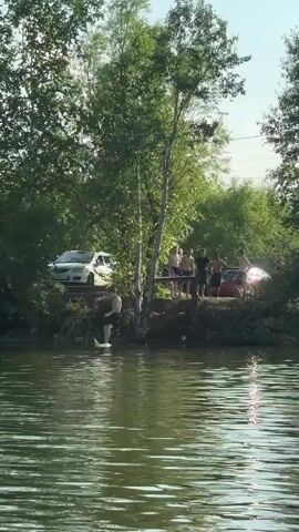A group of people are standing on the bank of a body of water, with trees and vegetation behind them. A white car is parked on a dirt path to the left of the group, and a red car is parked to the right. One person from the group is in the water, creating a splash. The water appears to be murky green. The sky is visible through the trees and is a light blue with some clouds. The overall scene suggests an outdoor recreational activity.