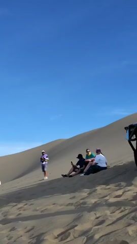 The video depicts a group of people in a desert landscape. Initially, a group is seen sitting on the side of a sand dune, with one person standing and holding a camera. Then, a yellow dune buggy is parked nearby, and several people gather around it. The camera pans out to show the vast desert landscape, revealing more sand dunes and distant mountains under a cloudy sky. Another dune buggy is seen driving off in the distance.