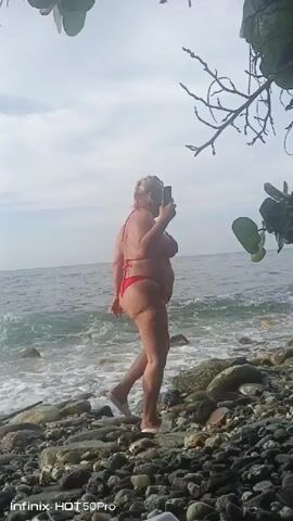 A woman in a bikini is walking on a rocky beach near the ocean. She is holding something in her hand and looking to her left. The waves are crashing on the shore. The sky is cloudy.