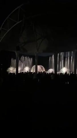 This video shows a nighttime outdoor performance featuring a water fountain show. The scene is framed by a dark, arched structure supported by columns. In the foreground, a large crowd of people are silhouetted against the illuminated performance area. The water fountains are lit with changing colors, starting with white and then transitioning to red. The water jets create various patterns and shapes, some appearing as tall columns and others as more dispersed sprays. At one point, circular patterns of water are visible in the center of the performance area. The lighting also creates splashes of yellow and green on the water. The audience members appear to be watching the show attentively, with some holding up phones to record the event. The overall atmosphere is one of a public spectacle.