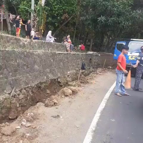 The video shows an outdoor scene with a damaged retaining wall next to a paved road. Several people are standing on the elevated area behind the wall, observing the situation. On the road, two young males are standing near an orange traffic cone, and a vehicle is partially visible. The environment appears to be rural or semi-rural, with vegetation in the background. The retaining wall has significant erosion and structural damage, with debris scattered on the road. The overall impression is of a recent or ongoing incident impacting the road and surrounding infrastructure.
