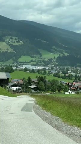 The video shows a sweeping panoramic view of a rural landscape with mountains in the background. In the foreground, there is a paved road curving to the left, with patches of water on it, suggesting recent rain. To the right of the road is a field of tall, dry grass. Further into the scene, there are clusters of houses with red roofs nestled amongst green rolling hills and trees. A small town or village is visible in the mid-ground, with buildings and what appear to be industrial structures. The mountains in the distance are covered in dense green forests, and the sky is overcast with clouds. The camera pans slowly from left to right, revealing more of the valley and the distant mountain range.
