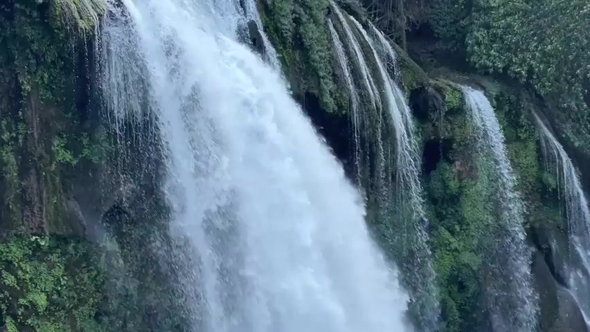 The video shows a waterfall cascading down a rocky, moss-covered cliff face. The water is white and foamy, with smaller streams trickling alongside the main flow. The environment appears lush and green.