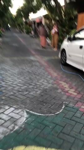 Two women and a baby are standing on a cobblestone street. The woman on the left is wearing a brown hijab and a peach-colored dress. The woman on the right is wearing a maroon hijab and a patterned yellow dress, and she is holding a baby. The street is lined with houses and lush greenery. There are colorful chalk lines drawn on the street, possibly indicating a game. A white car is parked on the right side of the street. The video appears to be filmed from a moving perspective, giving a blurred effect.