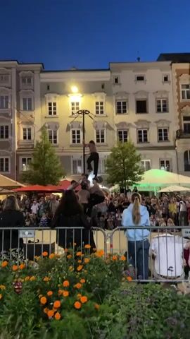 A group of performers is engaged in a human pyramid on a pole in a public square at dusk. A crowd of onlookers watches the performance. Buildings with lit windows form the backdrop. Trees and market stalls are also visible in the background.