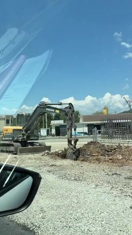 A yellow and black Volvo excavator is operating on a construction site. The excavator's arm is raised, and its bucket is digging into the dirt. The surrounding area is covered in gravel and dirt. In the background, there are buildings, trees, and mountains under a blue sky with white clouds. The video is filmed from a moving vehicle, as evidenced by the side mirror visible in the lower left corner.