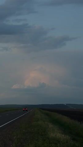 The media file shows a vertical shot of a road stretching into the distance, flanked by a grassy verge and a field on either side. A single car with its red taillights illuminated is driving away from the camera down the road. The sky is filled with dramatic, heavy clouds, suggesting an impending storm or the aftermath of one. The lighting indicates it is either early morning or late evening, with a soft, diffused light. The overall mood is somewhat somber and peaceful, with a sense of journey or passage. The landscape appears rural and vast, consistent with the location context of Kurgany, Russia. There is no visible text in the media file.