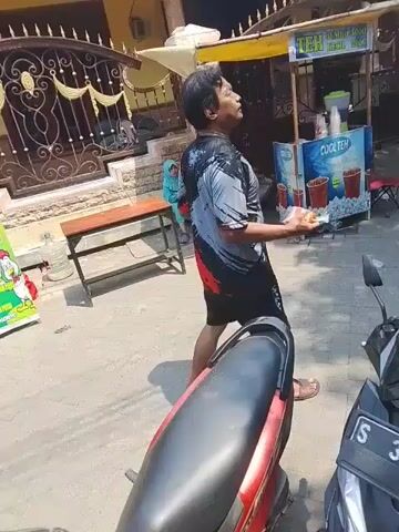 A busy street scene unfolds. In the foreground, a motorcycle is partially visible, its red seat and black handlebars indicating its presence. The camera pans to reveal a man wearing a black, red, and grey patterned t-shirt and black shorts, holding a dark object in his right hand and walking away from the camera. Behind him, two children, one in a light blue headscarf and the other in a white cap, are seated on a wooden bench. A wooden table is positioned to the left of the bench. Further back, a food stall with a colorful umbrella is visible, advertising "TEH COOL TEA". Several other stalls and people are present in the background, suggesting a market or roadside vendors. The environment appears to be outdoors with a bright, sunny sky.