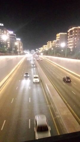 This media file depicts a nighttime scene of a multi-lane highway in Almaty, Kazakhstan. The camera perspective is elevated, looking down onto the road, which is well-lit by streetlights and the headlights of passing vehicles. The highway appears to be in an urban or suburban area, with modern buildings and illuminated facades visible on the left side of the frame. These buildings suggest a developed cityscape, potentially commercial or residential complexes.

Several cars are in motion on the highway, traveling in the same direction. The streaks of light from their headlights and taillights indicate movement, with some vehicles appearing as blurs. The road surface appears wet, reflecting the lights and suggesting recent rain or damp conditions, which could imply a humid or slightly overcast night. The sky is dark, consistent with nighttime.

The activity is the continuous flow of traffic on a highway at night. There are no visible people in the immediate foreground or on the road, but the presence of vehicles implies human activity and transportation. The overall mood is one of urban nocturnal transit, with the bright lights creating a dynamic and somewhat busy atmosphere despite the darkness. No text is visible within the frame.