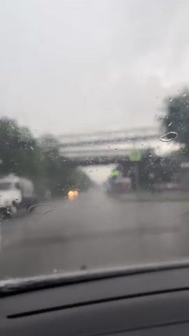 The video shows a view from inside a car on a rainy day. The windshield is wet with raindrops and streaks. Outside the car, it is raining heavily, and the road is wet and reflects the headlights of passing cars. There is a pedestrian overpass visible in the distance, as well as trees lining the road. Traffic lights and street signs are also visible. The camera is stationary, and the focus is on the scene outside the car. The overall impression is one of a dreary, wet day.