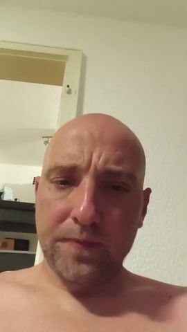 The media is a close-up selfie-style video of a bald man with stubble. He is indoors, with a slightly textured white wall and a doorway visible in the background. A shelf with some items is also visible on the left. The man's expression is somewhat serious, and he occasionally touches his head and neck. The lighting suggests it could be daytime or evening. There is no visible text in the media.