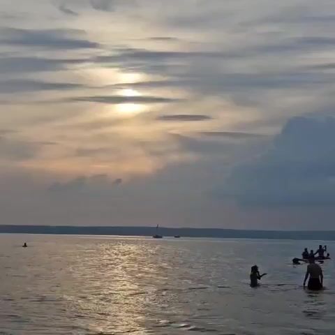 The video captures a scenic view of a body of water at dusk. The sky is a gradient of soft grays and blues, with the sun partially obscured by clouds, casting a gentle glow on the water. Several sailboats are visible in the distance, their sails catching the fading light. Closer to the shore, a few people are enjoying the water; some are swimming, while others are in a canoe. The overall atmosphere is serene and peaceful, with the natural beauty of the landscape taking center stage.