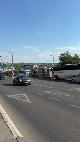 The video captures a street scene from the perspective of someone in a vehicle. The camera pans slightly to the right, showing a road with multiple lanes and traffic. Various vehicles are visible, including cars and buses. In the foreground, a black car is on the left side of the road. To the right, a large white and yellow bus is parked or moving slowly. Further down the road, more vehicles and pedestrians can be seen. There are buildings and trees lining the street, and the sky is clear and blue. The scene appears to be in an urban environment during daylight.