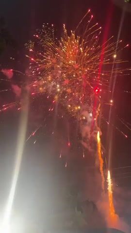 The video shows a night sky filled with fireworks. The fireworks are bursting in various colors, including orange, yellow, and red, creating bright trails and sparks against the dark background. Some fireworks are large and spherical, while others are long and streaky. There is a significant amount of smoke and haze in the air, which is illuminated by the light from the fireworks. In the lower left corner of the frame, a bright, out-of-focus light source is visible, possibly a flashlight or another light. The scene appears to be an outdoor event, such as a celebration or festival.