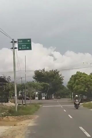 The video shows a road with a sign indicating directions to NISAM and BULOH BLANG ARA. A dark-colored car turns from a side road onto the main road, then proceeds down the main road. Trees and some buildings are visible on the sides of the road. The sky is cloudy.