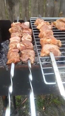 The video shows skewers of raw meat being grilled over hot coals. The scene is outdoors, with a wooden fence visible in the background. The meat appears to be chicken or pork, cut into bite-sized pieces and threaded onto metal skewers. Some of the meat is seasoned with spices. The grilling rack is made of metal, and there are visible embers beneath it. The overall impression is a casual, outdoor cooking setting, likely a barbecue or cookout. There is no visible text in the video. The time of day is not explicitly stated, but the lighting suggests it could be daytime. The weather appears to be clear. The location cues are limited to the fence, which is common in many outdoor settings. No people are visible in the video. The activity is clearly the preparation of food through grilling.
