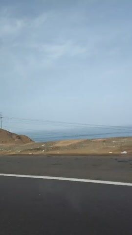 The video shows a view from a vehicle traveling along a coastal highway. The scene includes a paved road with a white line marking the lane, a strip of sandy or rocky ground between the road and the ocean, and the ocean itself. The sky is mostly clear with some scattered clouds. 

The road is mostly level, and the terrain adjacent to the road varies from sandy hills to rocky cliffs. There are utility poles with power lines running along the roadside. In one shot, the word "Jesus" is written on a hillside. The ocean is visible throughout the video, with waves crashing against the shore.  The camera appears to be mounted inside a moving vehicle, capturing the passing scenery.