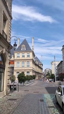 The video shows a street view on a sunny day. A light-colored building with a dark, mansard roof and several chimneys stands in the middle ground. A row of trees in planters lines the street in front of the building. People are walking on the sidewalk on the right side of the street, and one person is riding a bicycle. The street is paved with cobblestones. A white car is parked on the right side of the frame, partially obscuring the view. The sky is blue with scattered clouds.