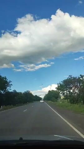 The video shows a view from the front of a car driving down a paved road lined with trees on both sides. The sky is blue with white clouds. A white car is visible ahead on the road.