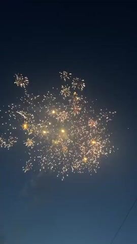 A slow motion close up shot captures a dynamic fireworks display against a dark blue evening sky. A bright green firework is seen firing from the bottom left, illuminating the left side of the frame with a strong green glow. In the center, multiple fireworks explode sequentially, creating vibrant bursts of light in shades of gold, orange, and red, with sparks showering outwards. The explosions light up a small section of the sky, creating a contrast with the dark background. A faint, thin white line is visible in the upper right corner, possibly a contrail or a distant object. The overall scene is lively and celebratory, dominated by the brilliant light and movement of the fireworks.