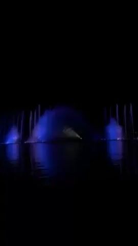 The video shows a water fountain at night, illuminated with alternating blue and purple lights. The fountain is located in a body of water, which reflects the colored lights. The fountain display features multiple vertical streams of water and a broader, arch-shaped spray in the center.