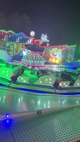 A nighttime amusement park ride named "DJ MACHINE" is in motion, illuminated by bright green and blue lights. The ride features a central spinning platform surrounded by multiple individual pods that also spin and move. On the backdrop of the ride, images of people, possibly DJs or performers, are visible. The overall atmosphere is festive and energetic, indicative of a carnival or fairground setting.