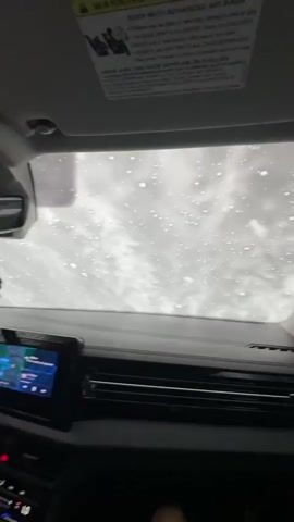 The video shows the interior of a car, primarily focusing on the view through the windshield and the front passenger window. The car is moving through an automatic car wash. Water and white foam or soap are sprayed across both the windshield and the passenger window. Large rotating brushes are visible through the passenger window, actively cleaning the car's exterior, causing the water and foam to swirl and streak across the glass. The car's dashboard, air vents, and a sun visor are visible inside the vehicle.