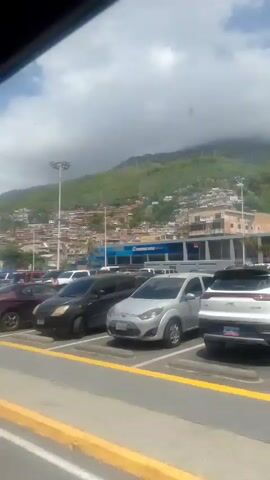 The video shows a street scene with several parked cars and buildings. A blue car is parked in the foreground on the left. Behind it, a white pickup truck is driving. Further back, there are several other cars parked. In the background, there are buildings with signs, including one that says "MYKONOS" and another that says "CENTRO". There are also hills with houses built on them in the distance. The sky is cloudy. The video pans from left to right, showing different parts of the scene. At one point, a person wearing a blue shirt and jeans is visible walking in the middle of the street. Another person, wearing a pink shirt, is visible on the right side of the screen. The video ends by showing more buildings and cars.