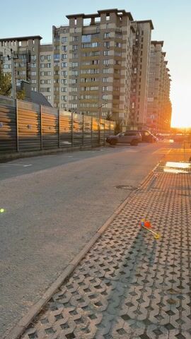 A short, static video reveals a street scene.  In the foreground, on the right side of the screen, a colorful child's toy lies on a patterned brick sidewalk. The street, made of asphalt, stretches into the distance where a line of modern, high-rise apartment buildings is visible. The buildings are bathed in the golden light of the setting sun.  The sun's rays are also reflected on a dark-colored fence running along the left side of the street. A dark-colored SUV is parked beside the fence. The sky above is a clear blue.  The camera remains stationary throughout the video.