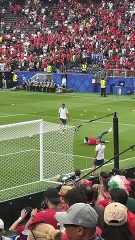 A soccer player in a red jersey is on the field near the goal. Another person in a white shirt stands behind him. A soccer ball is near the goal. There are spectators in the background wearing red and white clothing. The scene appears to be during a soccer match.