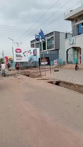 The video captures a street scene in Mubi, Nigeria, under an overcast sky, suggesting it might be morning or afternoon. The road is unpaved and shows signs of disrepair, with debris and a visible drain.

Several people are present, primarily moving along the street. On the left, a yellow motorized tricycle, likely a "keke napep," is partially visible, with a person riding a blue motorcycle in front of it. Further down the road, a person is riding a bicycle. A few other individuals are seen walking or standing.

Dominating the background is a large billboard advertising "TOGETHER FOR PROGRESS 2027." It features a portrait of a man, identified as "CHIEF MAURICE VUNOBOLKI," with the text "FOR GOVERNOR ADAMAWA STATE." An umbrella symbol is also prominent on the billboard, possibly representing a political party or campaign. Two flags, one green and one blue with a logo, are mounted on a pole above the billboard.

Adjacent to the billboard are buildings, one of which appears to be a shop with goods displayed outside. Signs are visible on the buildings, including "NYIONE GYS & CO. NIG. ENT." and "POS." A street lamp is also visible.

The overall impression is of a typical urban street in Nigeria, with the presence of political campaigning, local transportation, and commercial activity. The mood appears neutral, focusing on the everyday life and visual elements of the location.