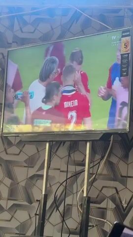 The video displays a television screen showing a soccer game. The game is being broadcast on TVRI. The screen shows players in red and blue uniforms interacting with each other. Some players are holding water bottles and appear to be taking a break during the game. One player in the foreground wears a red jersey with "SUISSE" written on the back. The background of the room where the TV is located is a patterned wallpaper. The TV is mounted on a stand.