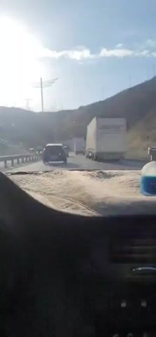 A perfume bottle sits on the dashboard of a car, facing forward. The car is in traffic, with other vehicles visible on the road ahead. The sun is shining, and the sky is blue with some clouds. The background shows a mountainous landscape.