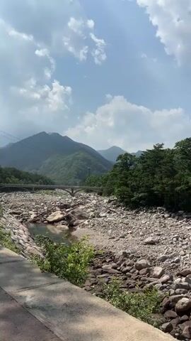 A scenic drone shot pans across a rocky riverbed dotted with various sizes of stones, with a narrow stream of water flowing on the left. Lush green mountains rise in the background, partially obscured by trees and a clear blue sky with scattered clouds. A bridge is visible in the distance. A paved path runs along the right side of the riverbed, with greenery growing along its edge. The camera's movement is smooth and steady, capturing the natural beauty of the landscape. There are no people or animals visible in the video.