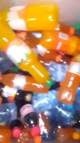 A close-up, slightly shaky video shows a pile of various plastic bottles. There are different colors, including orange, yellow, clear blue, and dark brown. The bottles appear to be various sizes and contain different beverages, including juices, water, and soda. Some of the bottles have labels visible. The background is blurry and indistinct.