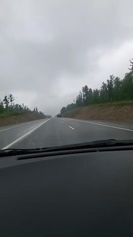The video, taken from the perspective of a driver inside a vehicle, shows a gray, overcast sky and a wet asphalt highway. Trees and a dirt embankment line the right side of the road, with sparse trees on the left. Several vehicles are visible on the highway in the distance, including a red car and two orange construction vehicles. The perspective is looking forward along the road, with the dashboard visible in the foreground.