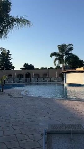 A wide shot shows an empty swimming pool area in the early morning or late afternoon. The sky is clear and blue. In the foreground, a paved patio with large, grey interlocking stones is visible, with a lounge chair at the bottom right. To the left, a palm frond hangs into the frame. Beyond the patio, a swimming pool with clear blue water is seen. Several lounge chairs with white cushions are lined up behind the pool, shaded by umbrellas. A beige building with arched openings runs along the back of the pool area. A palm tree stands to the right of the building, its fronds silhouetted against the sky. A small bar area with a white roll-down shutter is located on the right side of the pool. The overall impression is one of tranquility and emptiness, suggesting the area is not yet open for use.