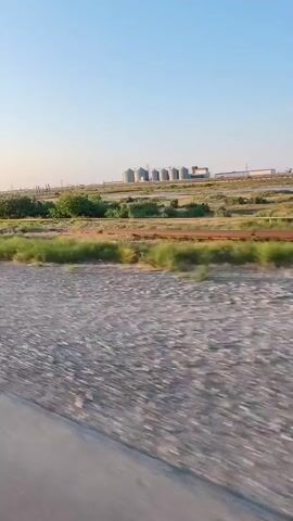 The video shows a view from a moving vehicle, possibly a car or train. The foreground consists of a gravelly surface, likely a road or track. In the middle ground, there is a stretch of dry vegetation with some green bushes and trees scattered throughout. In the background, a row of industrial silos, painted in muted colors like white and gray, are visible against a clear, pale blue sky. The overall scene appears to be an agricultural or industrial landscape in a dry climate, likely during daylight hours given the brightness and clear sky. The camera's perspective suggests the viewer is traveling parallel to the silos.