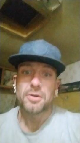 Certainly! Here's a detailed description of the video:

**Scene Description**

The video features a close-up shot of a man indoors. He is wearing a light gray t-shirt and a dark blue baseball cap with a flat brim. The cap has a subtle pattern. He also sports a nose ring and appears to have stretched earlobes with earrings that resemble a number "1."

The man has a light beard and mustache. The background is somewhat blurry, but a wooden frame is visible on the wall. A skylight or some form of window is present in the ceiling.

**Actions/Interactions**

The man is speaking directly to the camera, pointing his finger at the viewer at first. He then rubs his forehead with his right hand. He ends by smiling slightly.

**Environment**

The setting appears to be inside a room, possibly a home or trailer, based on the interior décor and lighting.