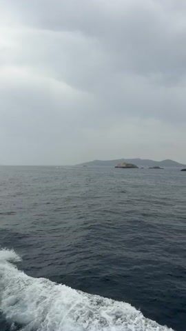 A boat is traveling across a dark body of water under a cloudy sky. The wake of the boat is visible in the lower left of the frame. In the distance, there are several islands. The closest island is small and rocky, with some vegetation. The other islands are larger and appear to have hills. The sky is overcast with gray clouds. The water is dark blue and choppy.