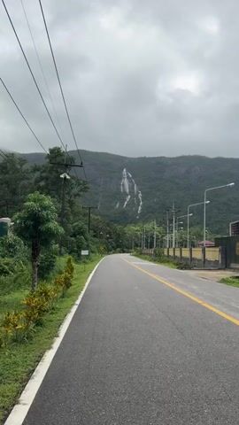 The video shows a paved road with a yellow line down the middle, curving to the left. The road is flanked by green vegetation and trees. Power lines are visible overhead. In the distance, a lush green mountainside is visible with several waterfalls cascading down it. The sky is overcast. A few streetlights are visible along the right side of the road.
