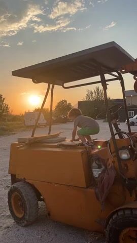 A young boy climbs on top of an old orange forklift at sunset. The forklift is parked in a lot with other vehicles and buildings visible in the background. The sun is setting behind the forklift, casting a warm glow on the scene. The boy is wearing a gray shirt and green shorts. He appears to be exploring the forklift.