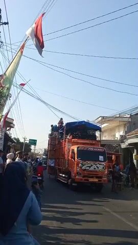A bright orange truck with large speakers and people on top is moving down a street. The truck has "GAB" written on the front. There are many people on the sides of the street, watching the truck. Red and white flags are hanging from wires above the street. The sky is clear and blue.